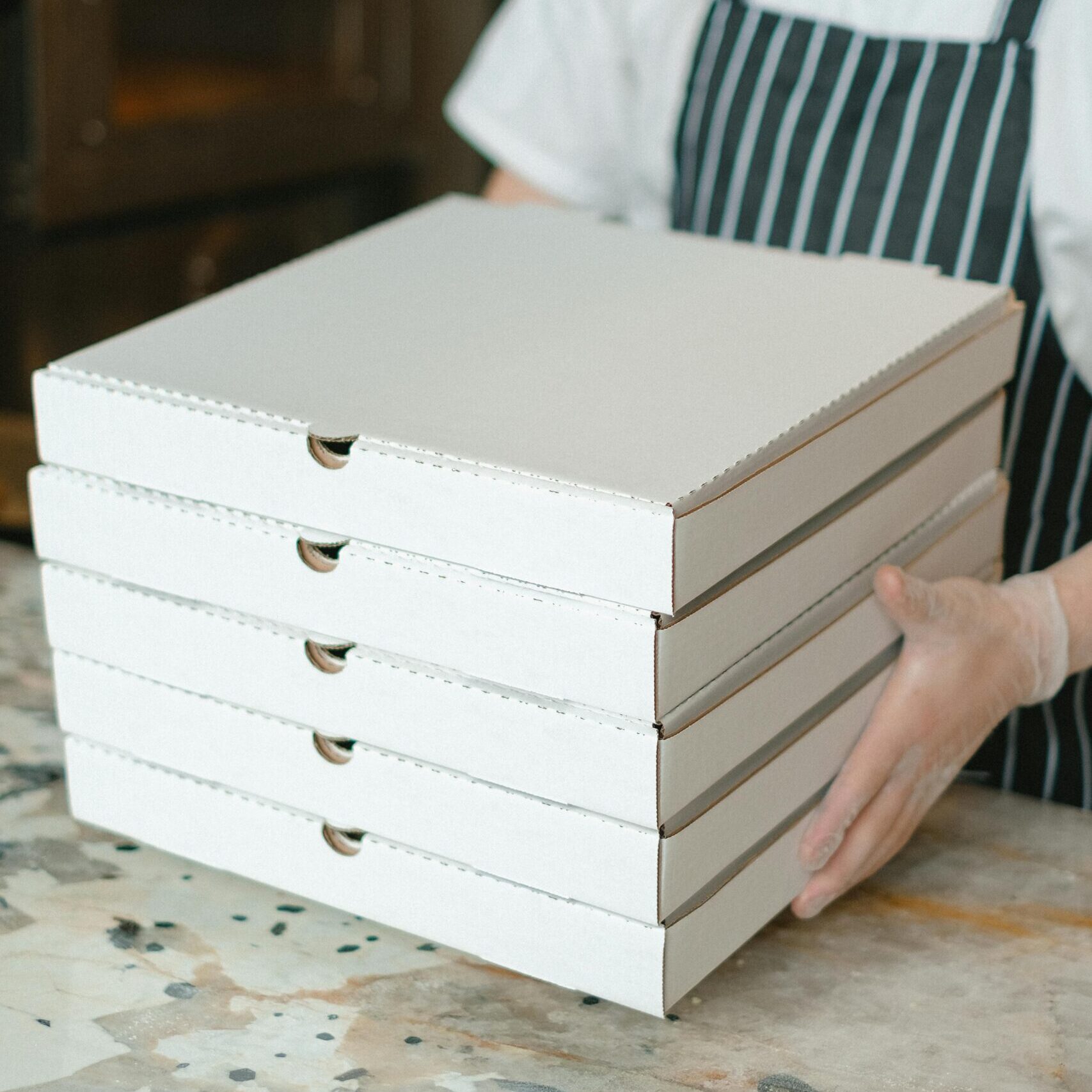 A pizza delivery worker holds stacked boxes in a restaurant kitchen, ready for delivery.